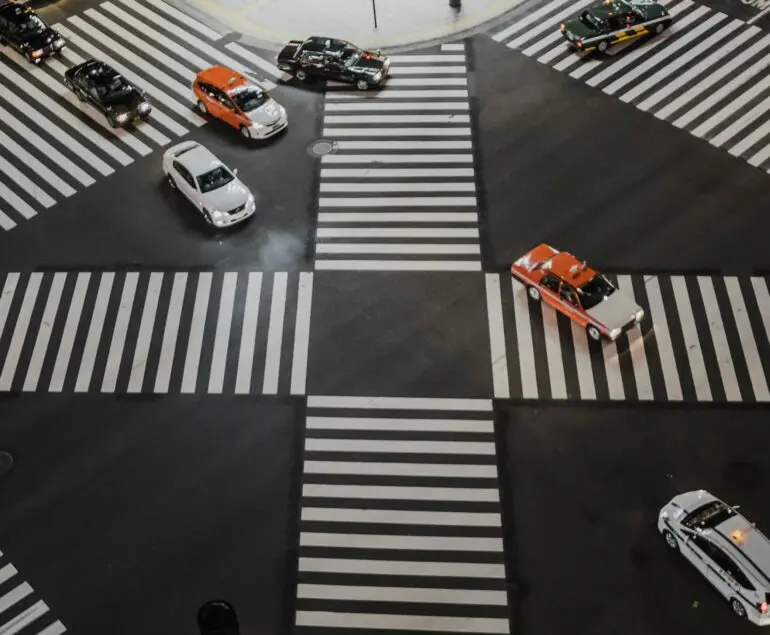 Aerial view of a busy Japanese intersection with multiple cars, including taxis, moving in different directions across a crosswalk.
