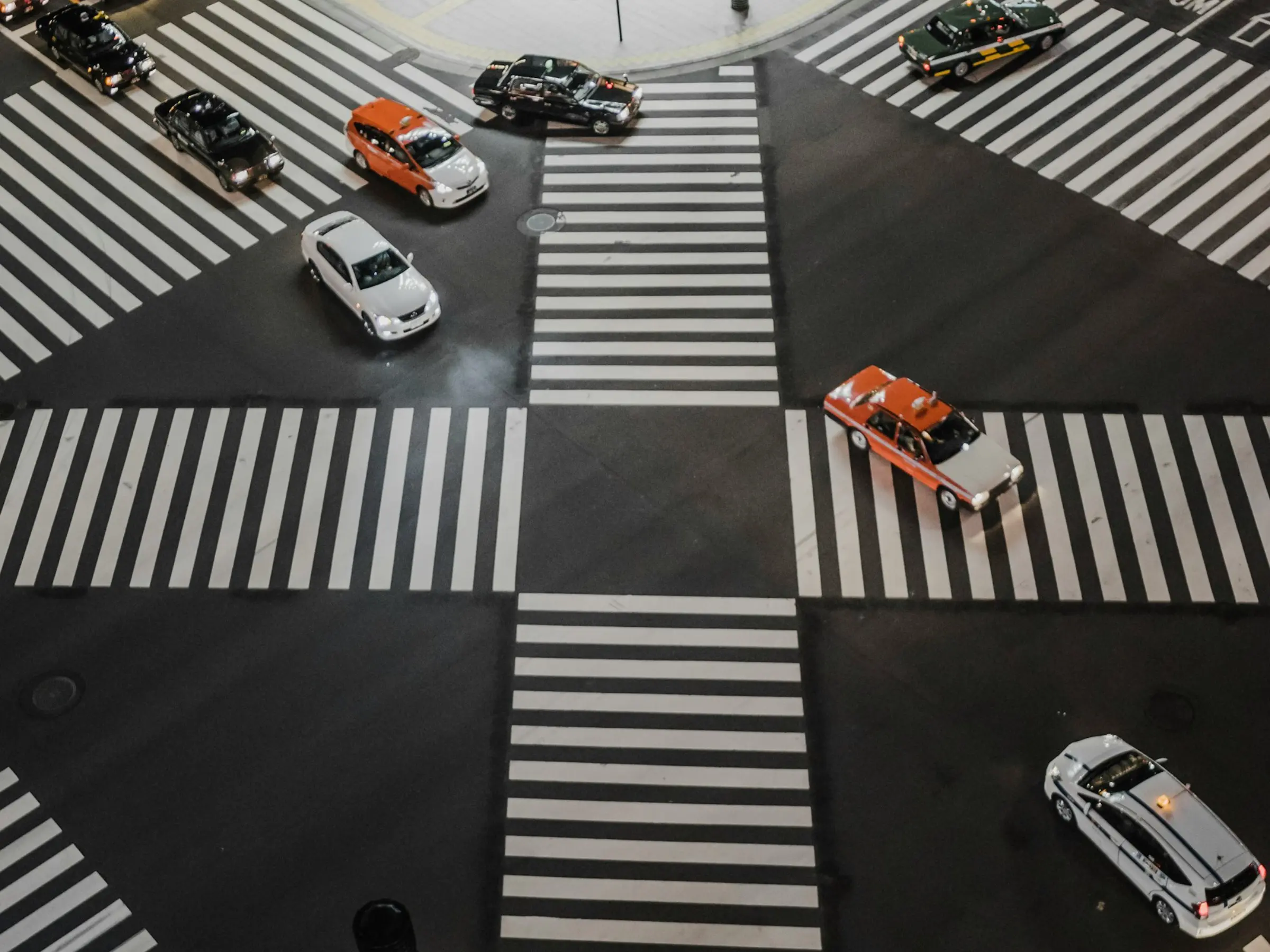 Aerial view of a busy Japanese intersection with multiple cars, including taxis, moving in different directions across a crosswalk.