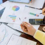 A person in a yellow shirt holding a pen over pages with graphs on a desk.