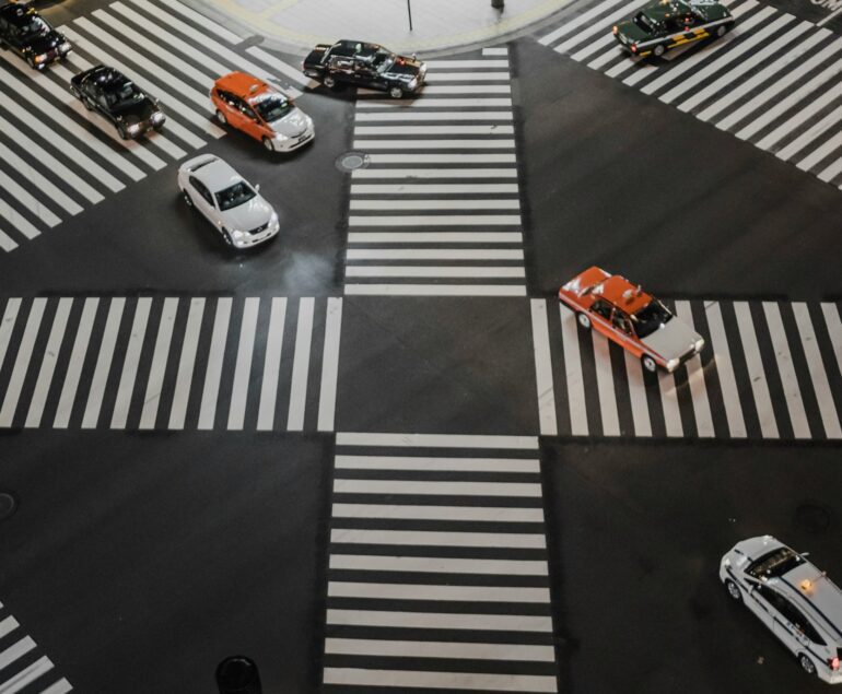 Aerial view of a busy Japanese intersection with multiple cars, including taxis, moving in different directions across a crosswalk.
