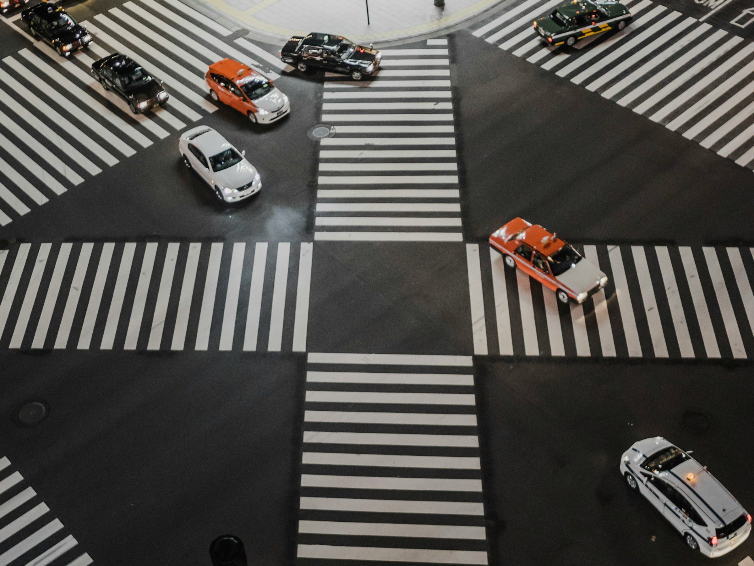Aerial view of a busy Japanese intersection with multiple cars, including taxis, moving in different directions across a crosswalk.