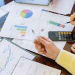 A person in a yellow shirt holding a pen over pages with graphs on a desk.